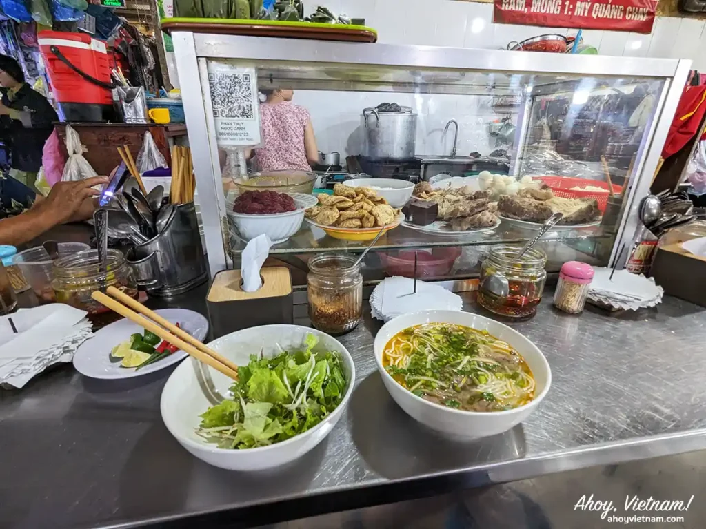 Bun bo in the My An Market in Da Nang, Vietnam, showing the bowl of noodles and beef, lettuce, the shop owner, and other food available for purchase
