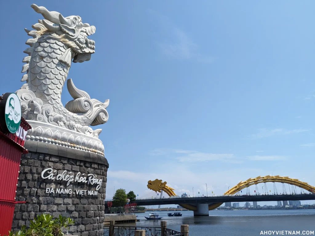 A dragon statue on the left and the yellow Dragon Bridge in the background on the right over the Han River in Da Nang, Vietnam