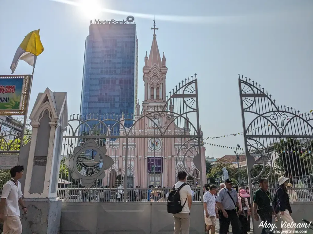 A sunny day outside the Da Nang Cathedral in Hai Chau, Da Nang, Vietnam, showing the tall, pink church next to an office building, with tourists entering into the church's grounds and taking photographs.