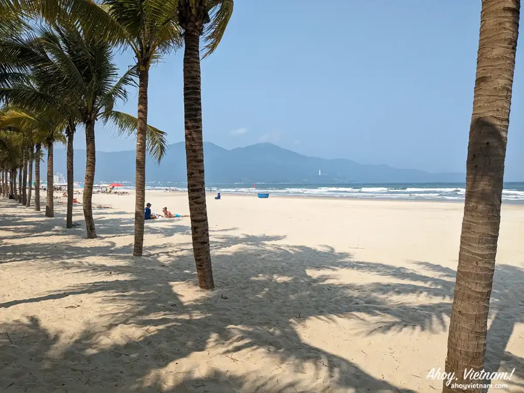 A sandy beach, palm trees, the ocean, the white Lady Buddha statue and Son Tra Peninsula in the background in Da Nang, Vietnam.