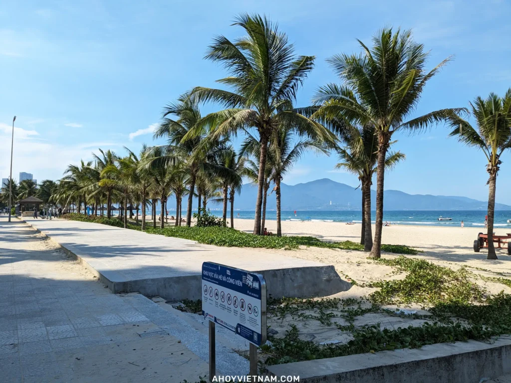 My An Beach in Da Nang, Vietnam, with sand, palm trees, tourists, Son Tra Mountain, and Lady Buddha in the distance.