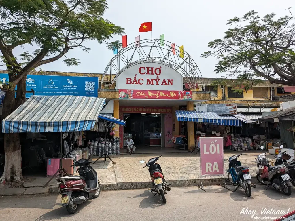 Outside the My An Market in My An, Da Nang, Vietnam, showing the market's entrance, parked motorbikes, signs in Vietnamese, and shopping booths