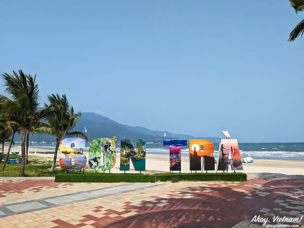 A multi-colored sign reading "Son Tra" with the beach behind it, palm trees, the Son Tra Peninsula, and the white Lady Buddha statue in the background in Da Nang, Vietnam