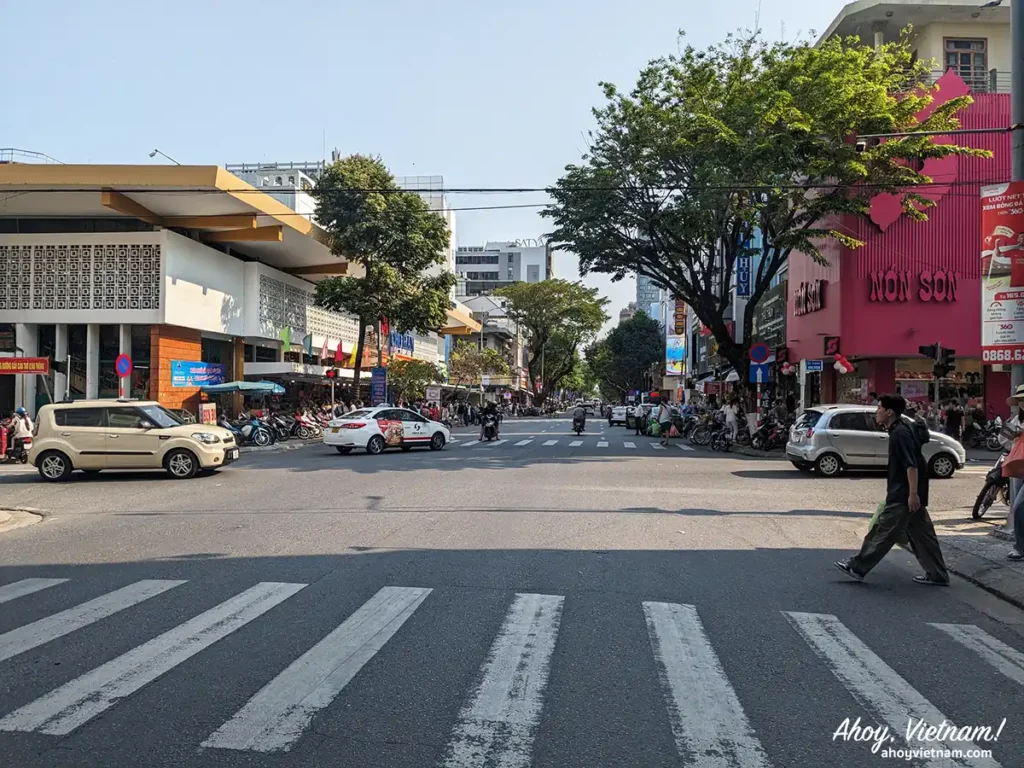A busy street in Hai Chau, Da Nang, showing pedestrians crossing the street, motorbikes, cars, the Han Market on the left-hand side, and shops on the right side.