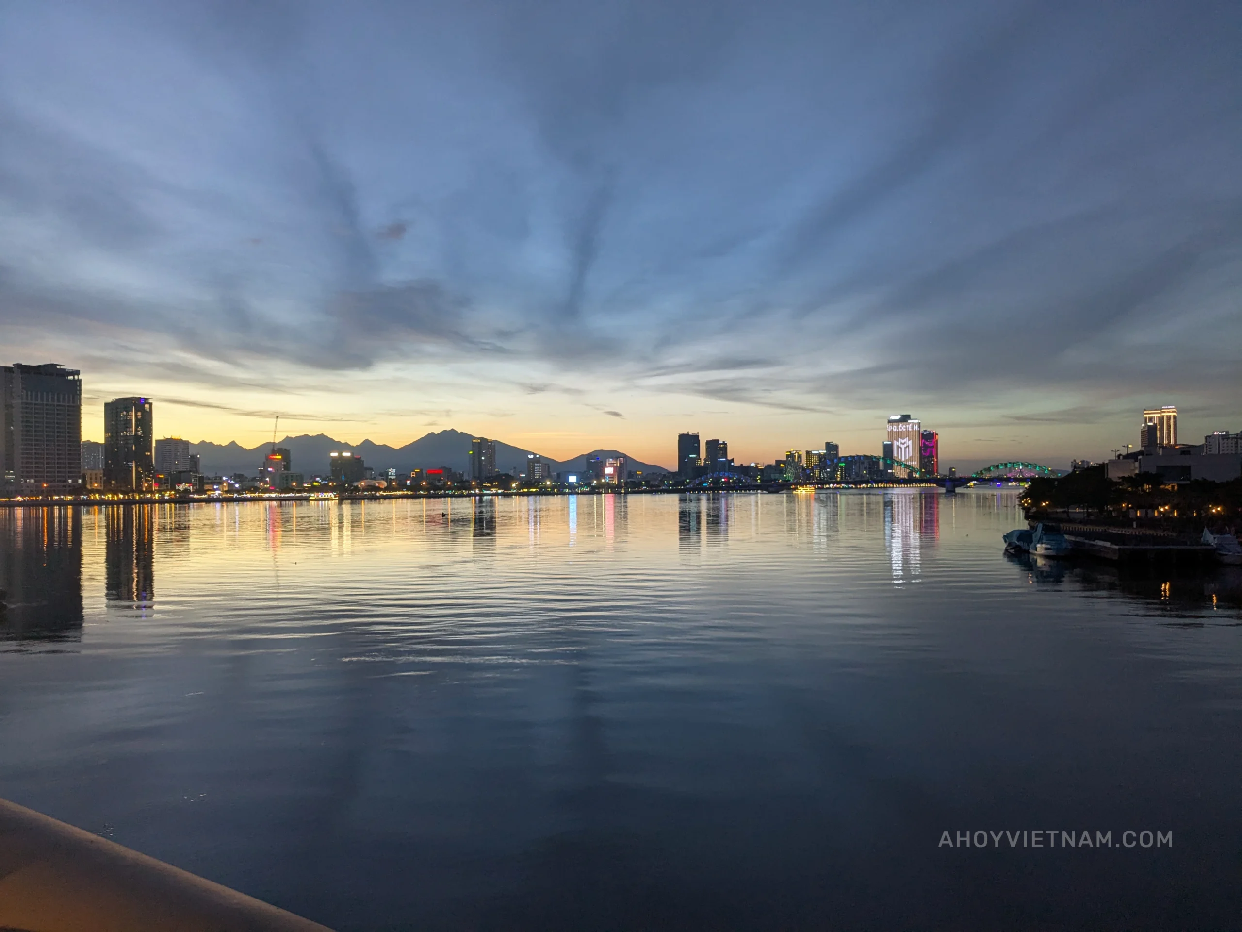 Sunset over the Han River in Da Nang, with mountains in the distance and buildings with neon lights.