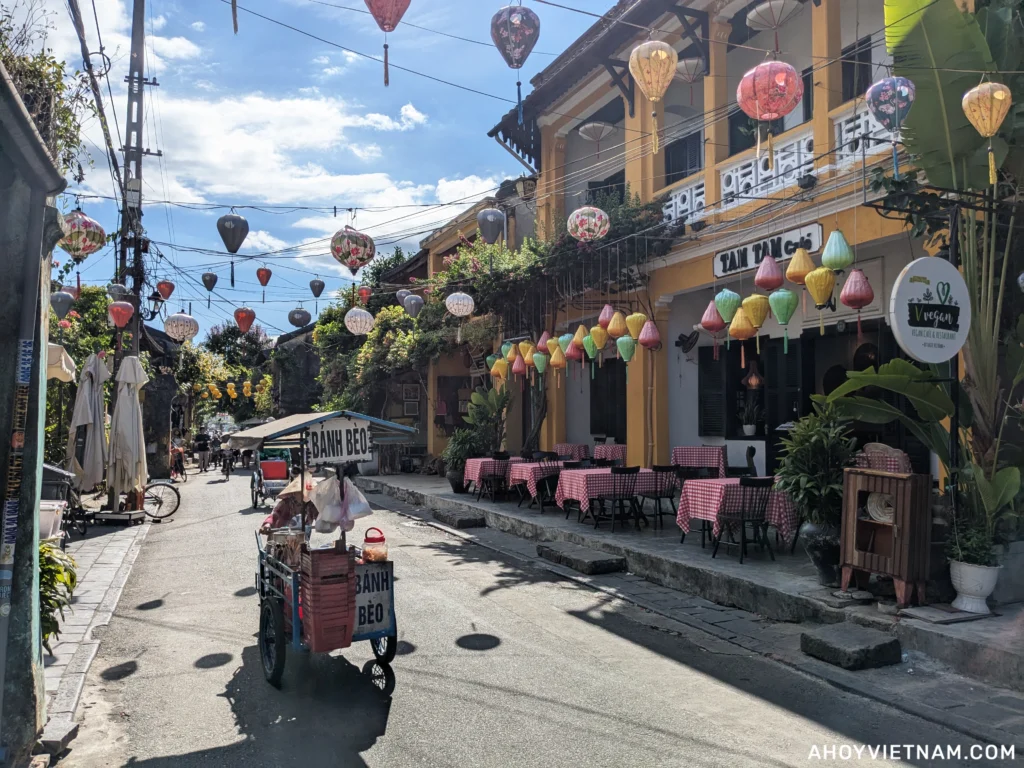 Hoi An Old Town, with a street vendor selling bánh bèo, lanterns overhead, and small restaurants lining the road.