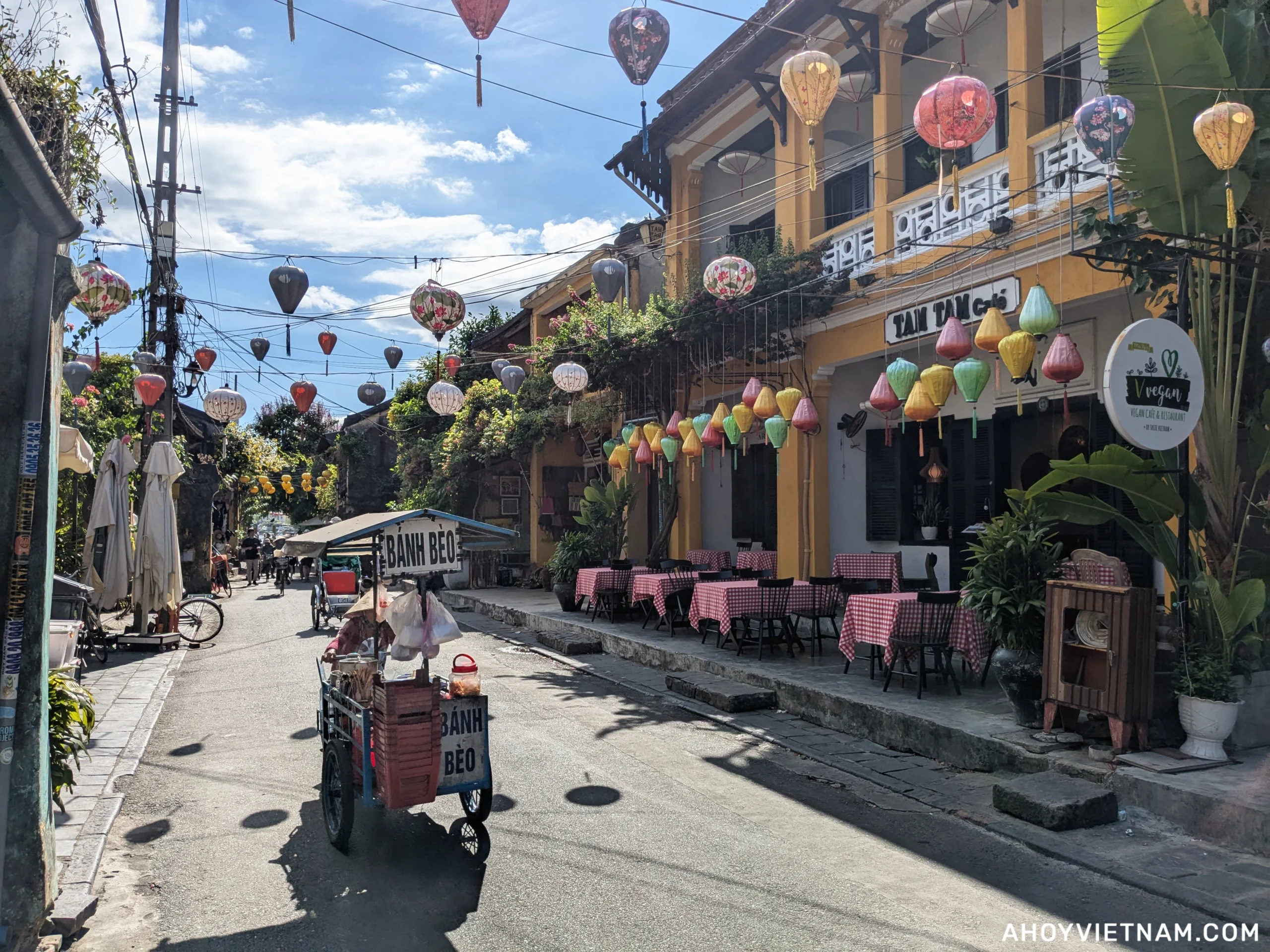 Hoi An Old Town, with a street vendor selling bánh bèo, lanterns overhead, and small restaurants lining the road.