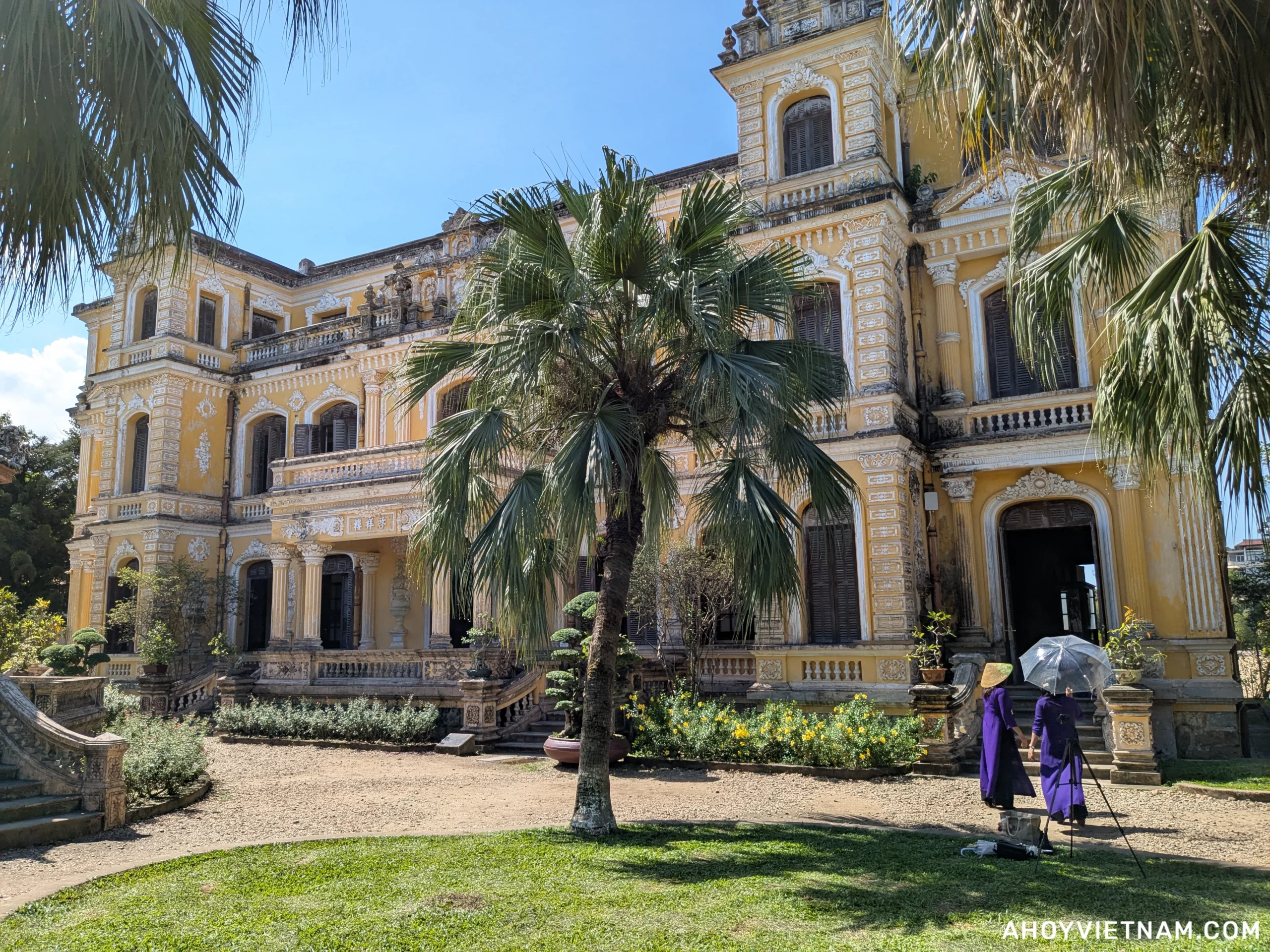 The front of An Dinh Palace in Hue, with two Vietnamese locals in purple Ao Dais and conical hats taking photos nearby