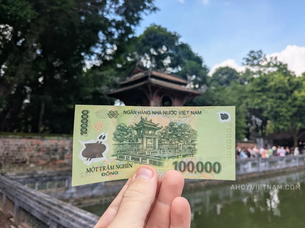 My hand holding up a 100.000 VND bill at the Temple of Literature in Hanoi, which is featured on the back of the bill