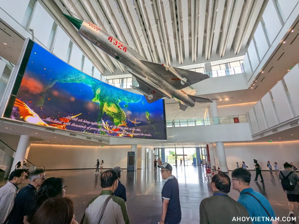 Visitors inside the Vietnam Military History Museum in Hanoi, with a large digital screen and an airplane hanging from the ceiling